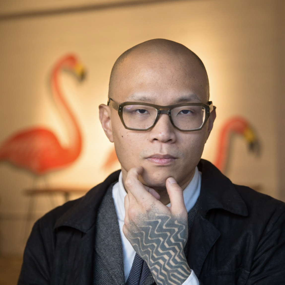 Photo of a bald man with glasses and hand tattoo with blurred flamingos in the background.